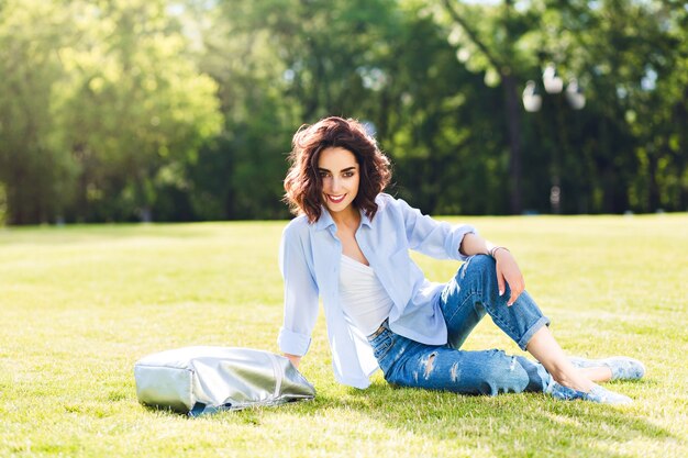 Foto de corpo inteiro de uma linda garota morena com cabelo curto, posando na grama na luz do sol no parque. Ela usa camiseta branca, camiseta e jeans, sapatos, bolsa. Ela está sorrindo para a câmera.