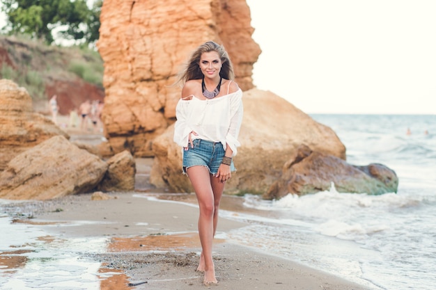 Foto de corpo inteiro de uma menina bonita loira com cabelo comprido, caminhando na praia perto do mar. Ela está sorrindo para a câmera.
