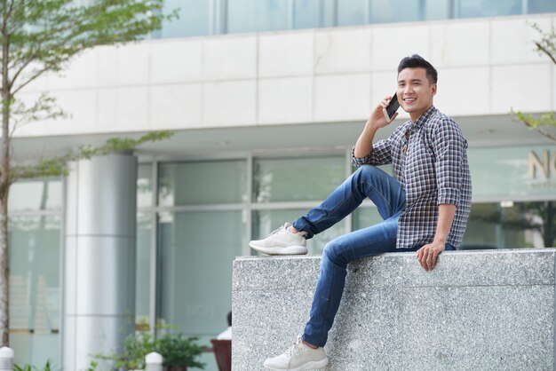 Foto de corpo inteiro do jovem asiático sentado no mármore ao ar livre, falando ao telefone