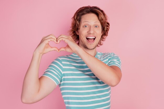 Foto de dedos afetuosos de homem louco mostrando o símbolo do coração e vestindo uma camiseta listrada azul casual com fundo rosa
