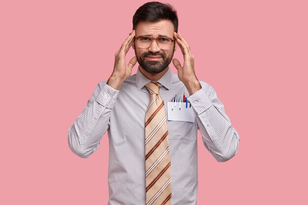 Foto de descontente homem com a barba por fazer sofre de dor de cabeça, mantém as mãos nas têmporas, tem expressão triste, cansaço depois do trabalho no escritório