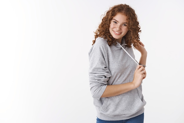 Foto de estúdio atrevida ruiva alegre carismática jovem vestindo um moletom cinza, sorrindo, boba, flertando, rindo, brincando com o cabelo, vestindo um moletom cinza casual, fundo branco em pé divertido sorte