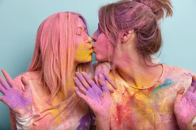 Foto de estúdio de duas jovens europeias satisfeitas se beijando, manchadas com o pó de holi colorido