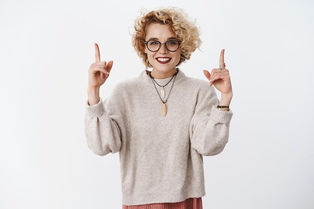 Foto de estúdio de elegante garota queer com batom de cabelo loiro curto e óculos apontando para cima com as mãos levantadas e sorriso entusiasmado, mostrando o espaço legal da cópia para promoção na parede branca.