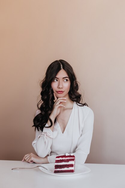 Foto de estúdio de encaracolado mulher asiática com sobremesa. Mulher japonesa pensativa posando com bolo.