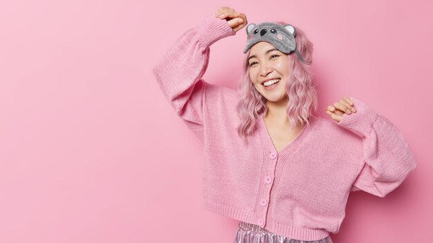 Foto de estúdio de feliz linda mulher asiática dança movimentos despreocupados com os braços levantados sente-se feliz por usar uma venda suave e um jumper casual isolado sobre o espaço em branco de fundo rosa para sua promoção