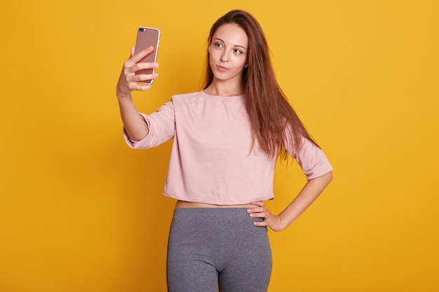 Foto de estúdio de linda mulher de cabelos castanho com cabelos lisos em calças cinza e camisa rosa tomando selfie