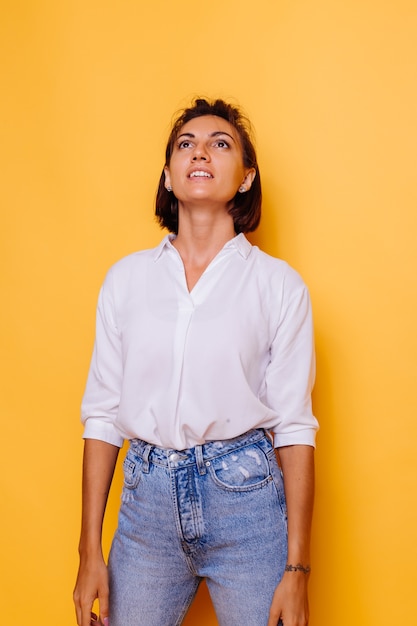 Foto de estúdio de mulher feliz, cabelo curto, vestindo camisa branca e calça jeans, posando na parede amarela