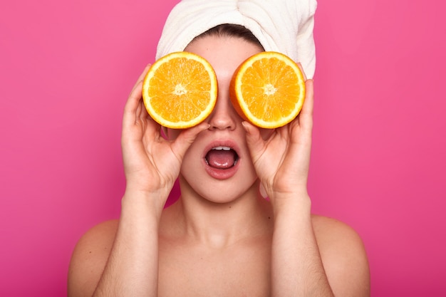 Foto de estúdio de mulher jovem e bonita chocado europeu agradável contras olho com laranjas, tem uma toalha branca na cabeça. modelo com pele clara posa em estúdio isolado na rosa. conceito de beleza.