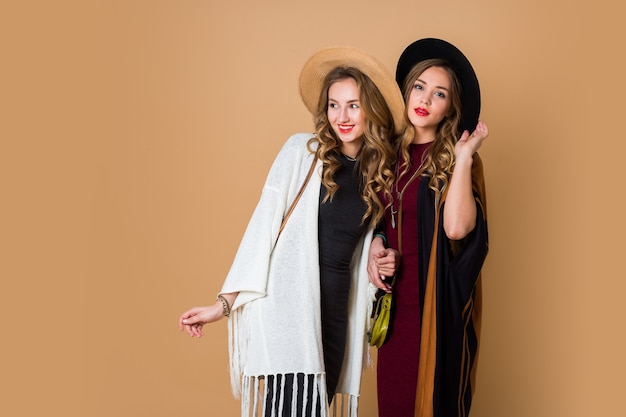 Foto de estúdio de outono de duas modelos com cabelo loiro ondulado em lã e chapéu de palha usando poncho listrado