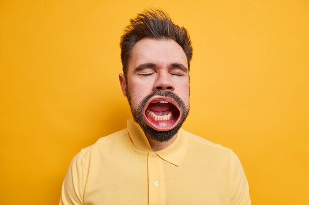 Foto de estúdio de um homem barbudo e cansado caucasiano fecha os olhos e mantém a boca bem aberta enquanto o bocejo mostra uma expressão sonolenta vestida com uma camisa