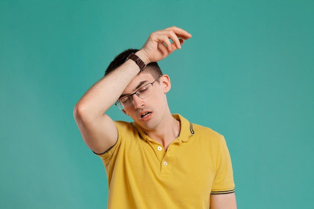 Foto grátis foto de estúdio de um jovem adorável em uma camiseta casual amarela, óculos e relógios pretos parecendo cansado enquanto posava sobre um fundo azul. corte de cabelo estiloso. conceito de emoções sinceras. copie o espaço.