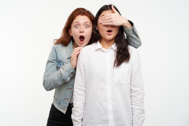 Foto de estúdio de um jovem casal interracial feminino, uma mulher estrelando a câmera com expressão facial chocada e fechar os olhos de outro com a palma da mão. isolado sobre fundo branco