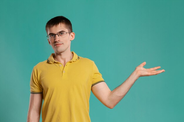 Foto de estúdio de um jovem elegante em uma camiseta casual amarela, óculos e relógios pretos agindo como segurando algo enquanto posava sobre um fundo azul. Corte de cabelo estiloso. Conceito de emoções sinceras