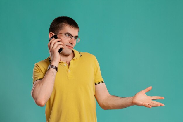 Foto de estúdio de um jovem inteligente em uma camiseta casual amarela, óculos e relógios pretos falando no celular e gesticulando enquanto posava sobre um fundo azul. Corte de cabelo estiloso. Emote sincero