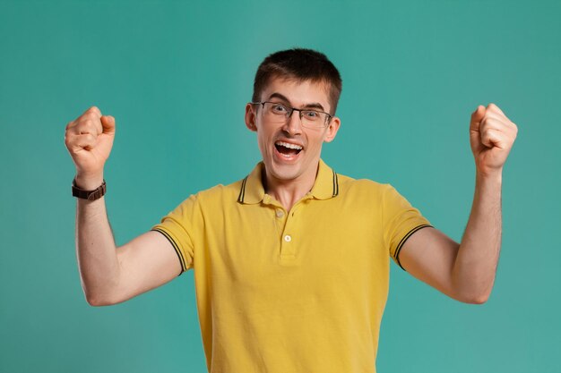 Foto de estúdio de um jovem interessante em uma camiseta casual amarela, óculos e relógios pretos parecendo feliz enquanto posava sobre um fundo azul. Corte de cabelo estiloso. Conceito de emoções sinceras. Copiar espaço