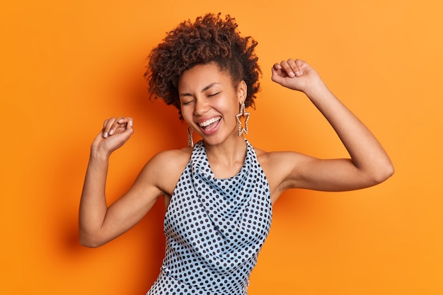 Foto de estúdio de uma bela jovem afro-americana despreocupada se movendo ativamente, levantando os braços e sentindo sorrisos enérgicos, gasta o tempo livre em uma festa discoteca isolada sobre o fundo laranja do estúdio