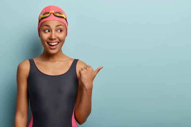 Foto de estúdio de uma feliz mulher de pele escura com um sorriso charmoso, usa trajes de banho e aponta para o lado com o polegar