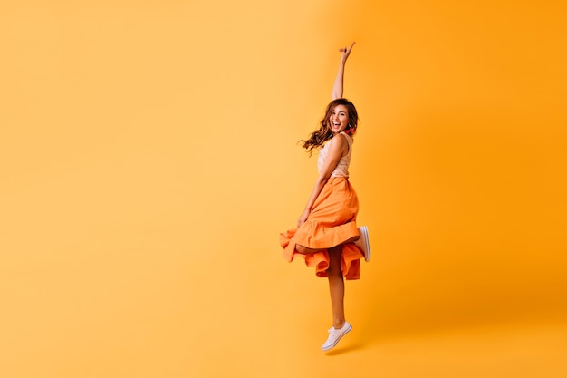 Foto de estúdio de uma garota bonita com saia laranja e sapatos brancos. Mulher ruiva animada pulando no amarelo.