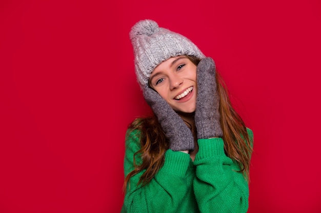 Foto de estúdio de uma garota feliz e sorridente com um sorriso adorável e olhos azuis tocando seu rosto, vestida de boné de inverno e luvas em fundo vermelho isolado