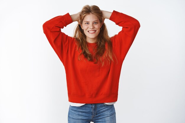 Foto de estúdio de uma jovem atraente e encantadora otimista com sardas fofas, sorrindo alegremente com um belo sorriso branco, segurando as mãos na parte de trás da cabeça em pose descontraída e descontraída, vestindo um suéter vermelho grande