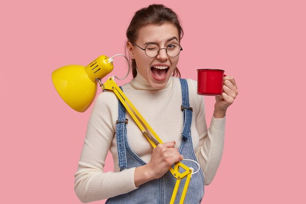 Foto de estúdio de uma linda mulher alegre pisca os olhos, se diverte com os amigos após fazer uma hometak, usa abajur, bebe chá quente ou café