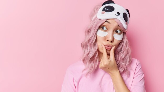 Foto de estúdio de uma linda mulher asiática faz beicinho olha de lado surpreendentemente aplica manchas de beleza sob os olhos vestidos casualmente isolados sobre o espaço de cópia de fundo rosa à esquerda para o seu anúncio