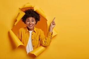 Foto de estúdio de uma mulher afro-americana positiva aponta o dedo para copiar o espaço acima, animada com boas informações, sorri agradavelmente, usa uma jaqueta amarela e fica em um buraco de papel rasgado.