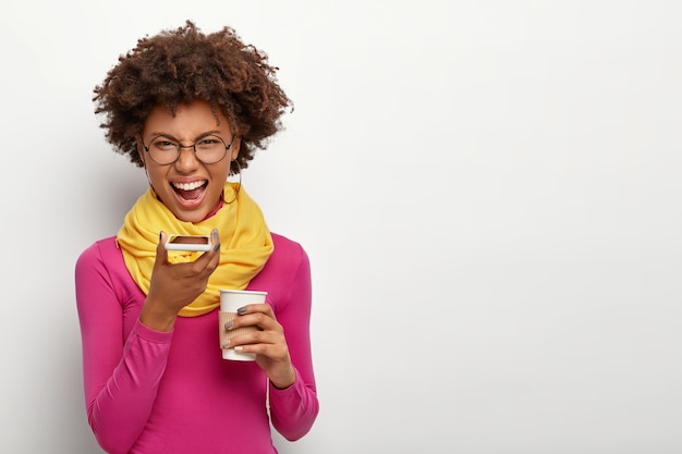 Foto de estúdio de uma mulher com raiva e irritada com penteado afro, faz chamada de voz via smartphone, bebe café para viagem, usa óculos óticos, blusa de gola alta rosa e lenço amarelo, isolado sobre a parede branca