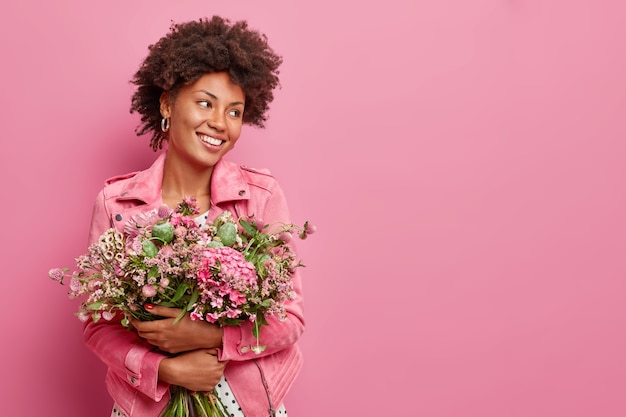 Foto de estúdio de uma mulher feliz segurando um grande buquê de flores celebra o feriado de primavera sorrisos alegremente desviando as poses contra a parede rosa com espaço de cópia para sua promoção