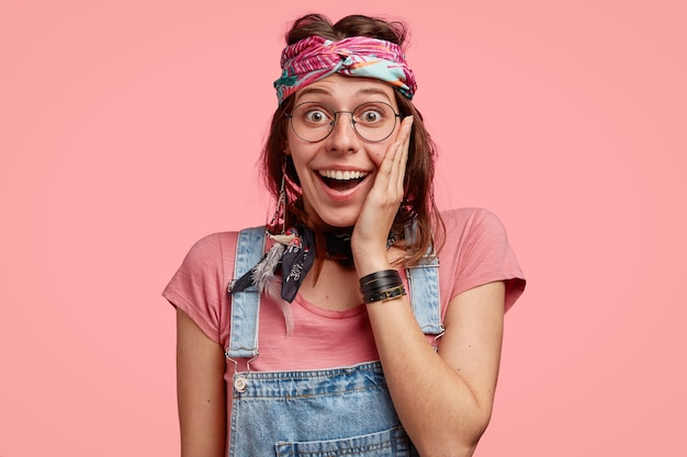 Foto de estúdio de uma mulher hippie estilosa com um sorriso largo, mão na bochecha e olhos arregalados