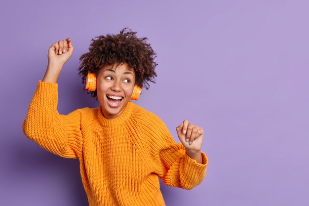 Foto de estúdio de uma mulher muito feliz se movendo com o ritmo da música levanta os braços e se sente otimista usando fones de ouvido estéreo jumper casual