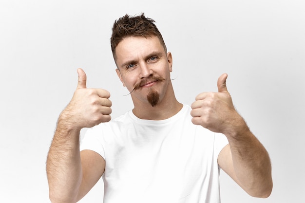 Foto grátis foto de estúdio isolada de bonito na moda jovem homem branco com cavanhaque e bigode de guidão olhando para a câmera com um sorriso amigável e positivo, mostrando o sinal de polegar para cima, gostando da ideia ou do plano