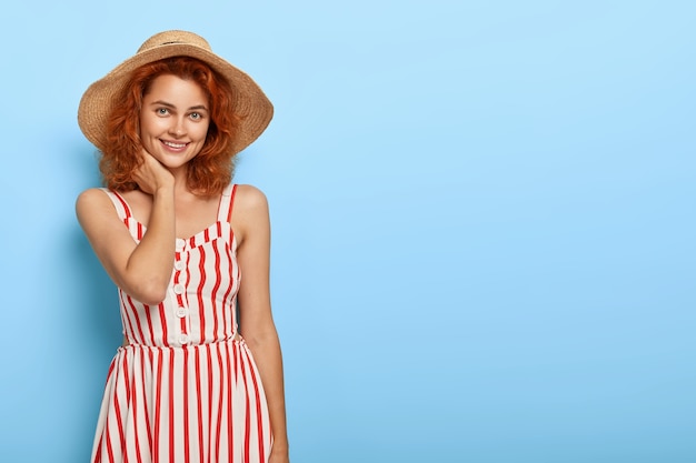 Foto de feliz mulher ruiva vestida com roupa da moda de verão, chapéu de palha, tem um sorriso encantador, beleza natural, fica contra a parede azul com espaço em branco para sua promoção.