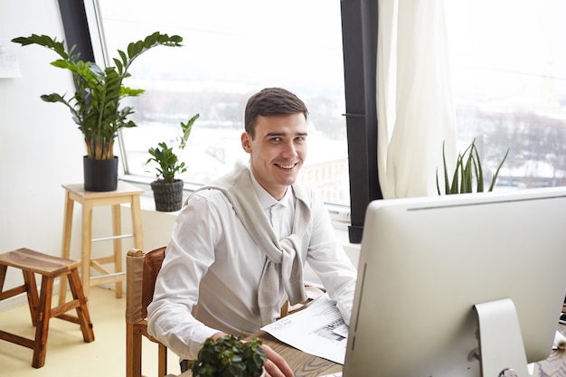 Foto de feliz sorridente jovem talentoso arquiteto masculino, projetando o projeto de construção em um escritório moderno, desenhando o plano e estudando as plantas na mesa, aproveitando o processo criativo. Trabalho e ocupação