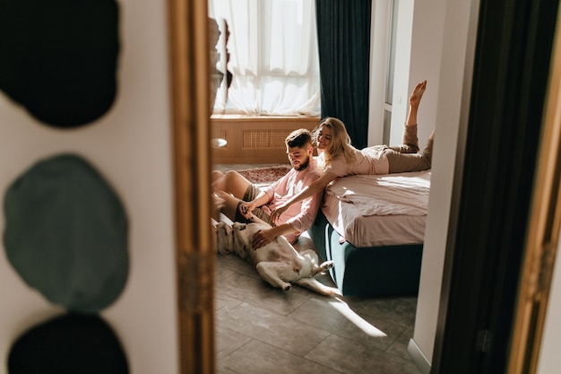 Foto de fim de semana de casal brincando com labrador no quarto ensolarado. garoto e garota estão acariciando a barriga de um cachorro grande.