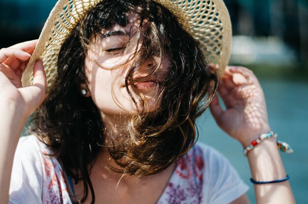 Foto de foco raso de uma mulher segurando seu chapéu enquanto aproveita o dia ensolarado com os olhos fechados