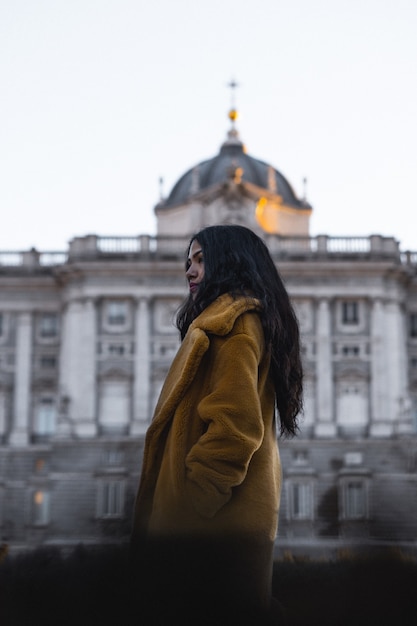 Foto de foco seletivo de uma jovem com um casaco marrom em frente a um ponto turístico da cidade