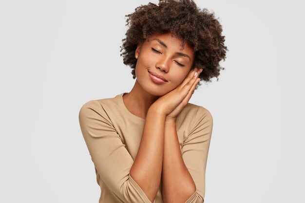 Foto de garota negra cansada cochilando, posa com as mãos juntas, mantém os olhos fechados, tem sonhos positivos, fica feliz em descansar