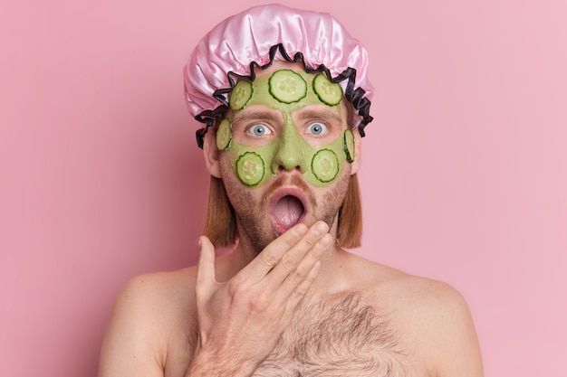 Foto grátis foto de homem chocado encara a câmera mantém a boca amplamente aberta aplica máscara de beleza facial para cuidados com a pele fica sem camisa interior contra um fundo rosa.