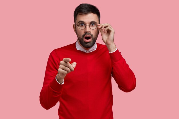 Foto de homem com a barba por fazer estupefato apontando diretamente com o dedo indicador, com expressão de medo, vestido com um macacão vermelho