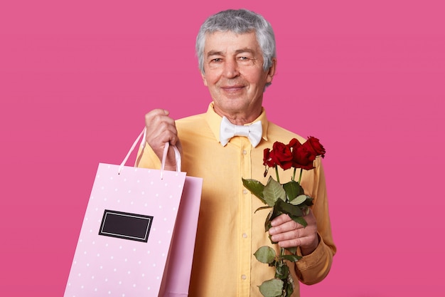 Foto de homem maduro atraente com expressão facial agradável, vestida de camisa amarela com gravata branca, carrega bolsa rosa com presente e rosas, quer parabenizar a esposa com o aniversário de casamento.