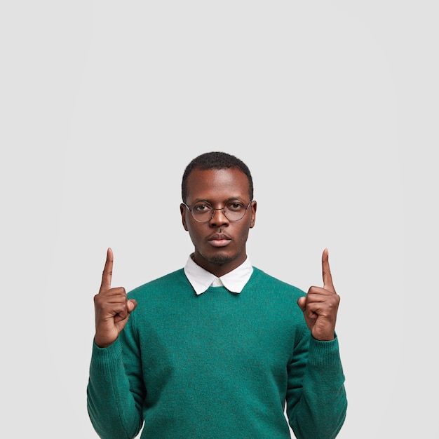 Foto de homem negro sério aponta com os dois dedos indicadores para cima, mostra o espaço livre acima, vestido com roupa estilosa