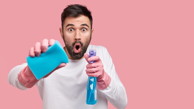 Foto de homem surpreso com a boca aberta, olhando em choque, carregando esponja e spray de limpeza, olhando estupefato, vestindo roupas brancas