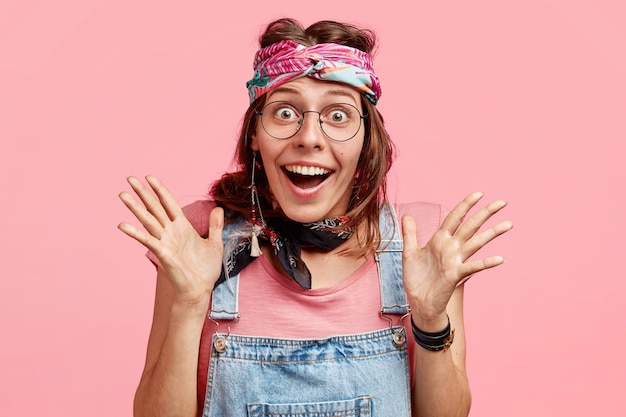 Foto de jovem alegre com expressão feliz, gesticula com as mãos, tem expressão radiante, não espera ver algo inacreditável, usa óculos e bandana, isolada no rosa