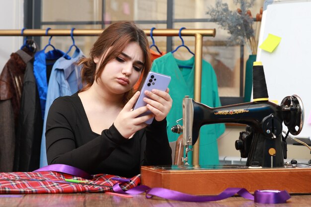 Foto de jovem alfaiate sentada à mesa e olhando para o telefone dela