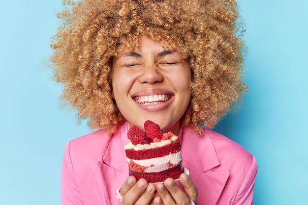 Foto grátis foto de jovem positiva com cabelo encaracolado segura bolo apetitoso com framboesas gosta de comer comida cremosa doce fecha os olhos de satisfação vestido com jaqueta formal rosa comendo sobremesa