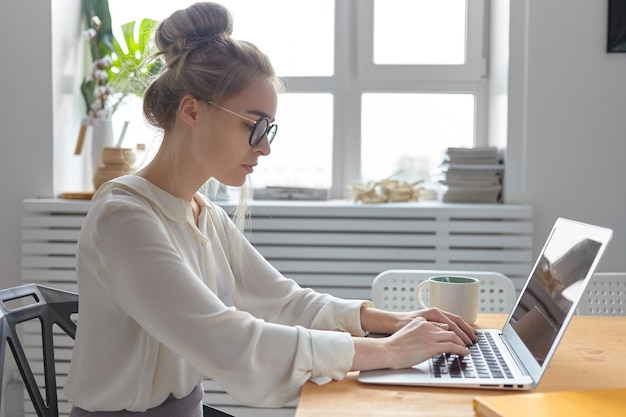Foto de lado de séria na moda jovem empresária europeia usando uma blusa branca elegante e óculos redondos digitando em um dispositivo eletrônico genérico, verificando e-mail, escrevendo carta comercial
