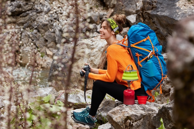 Foto de lado de um turista ativo fazendo uma pausa depois de passear, sentado na pedra, segurando uma câmera profissional