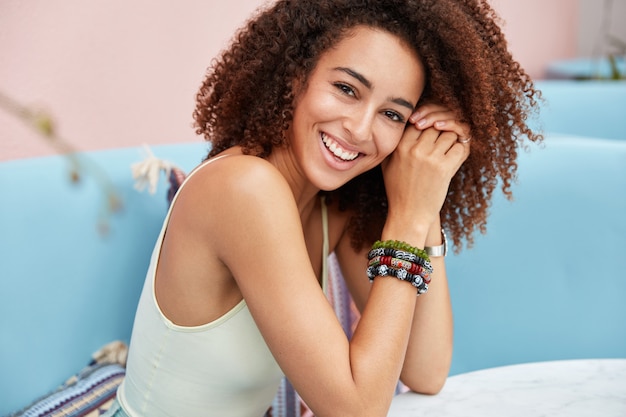 Foto de lado de uma jovem feliz, de pele escura, cabelos cacheados, sorriso largo e vestida com roupas casuais